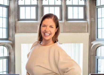 Kate Brown. A woman with shoulder length brown hair, smiling and standing in front of a stained glass window.