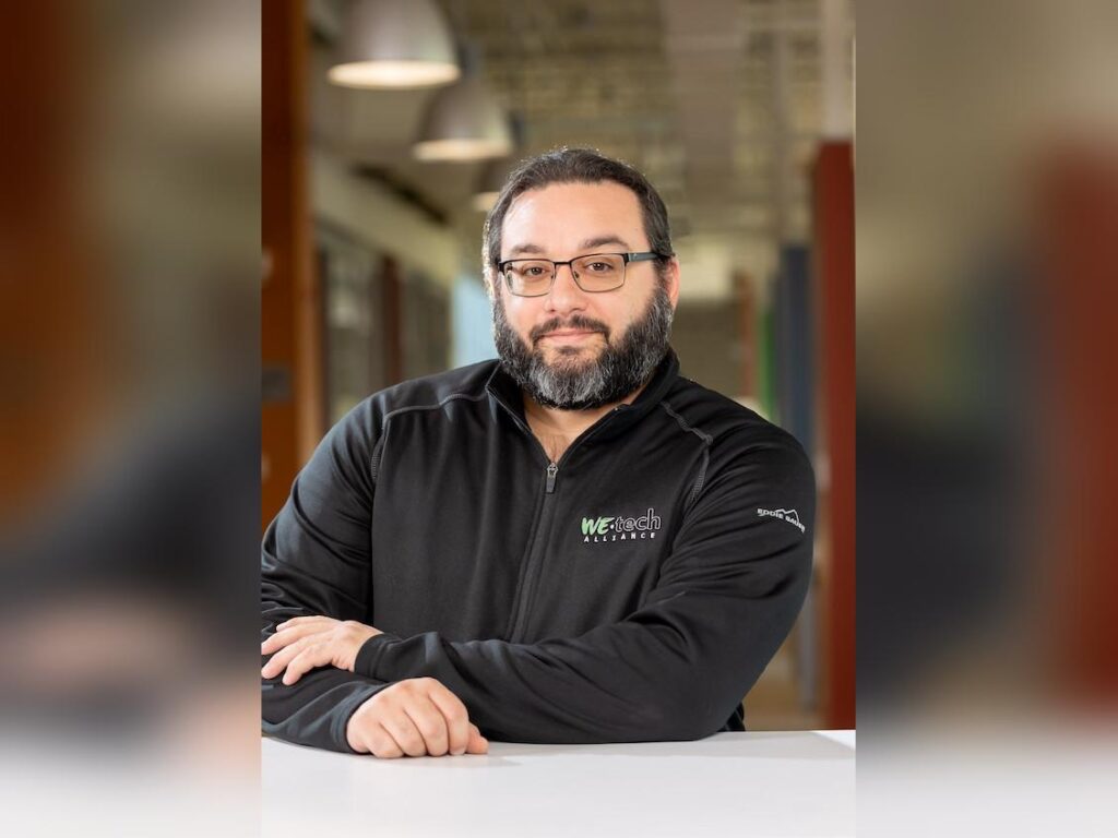 John-Marc is a gentleman with short, dark hair, glasses and a neat beard flecked with white.  He's wearing a sweater branded with the WEtech Alliance logo.