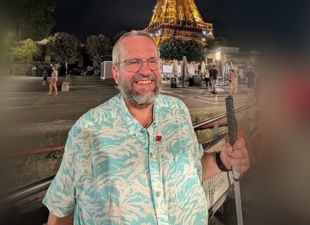 RIchard is a man with grey hair, moustache and bear. He is dressed in a colourful shirt and is wearing glasses and holding a white cane.