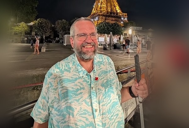 RIchard is a man with grey hair, moustache and bear. He is dressed in a colourful shirt and is wearing glasses and holding a white cane.